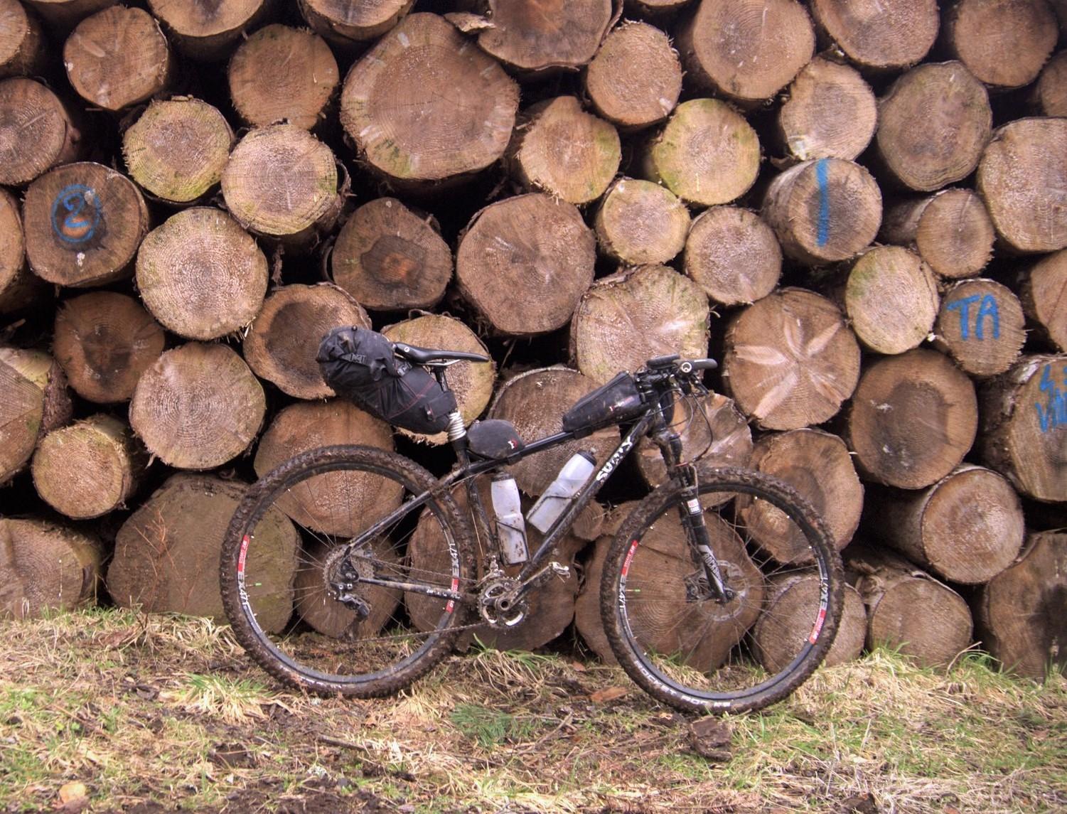 Surly Ogre: A mountain bike with a black saddlebag and a water bottle is leaning against a stack of large, round-cut tree logs. The ground is muddy, suggesting recent rain or wet conditions. The backdrop of logs adds a rustic, natural feel to the scene.