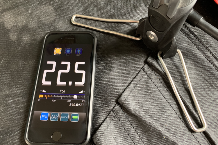 A smartphone displaying a reading of 22.5 PSI on a pressure gauge app, positioned next to a black fabric surface with a metallic tool and a clip nearby.