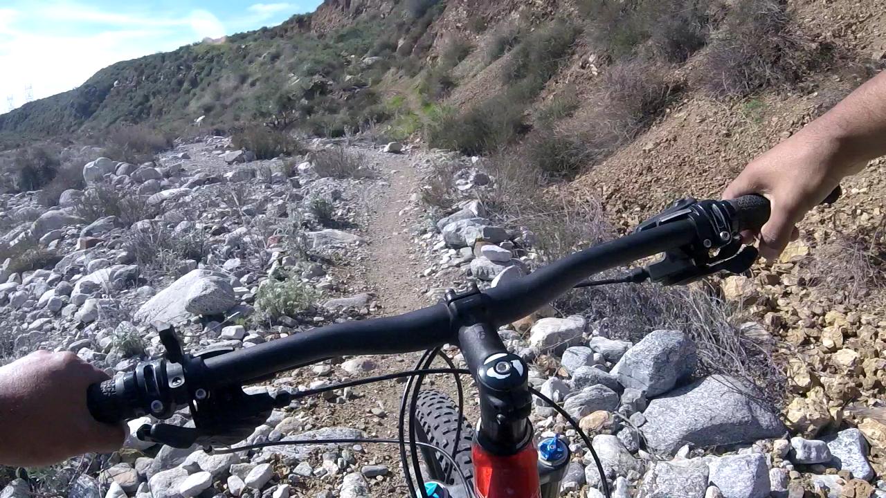 A first-person view of a mountain bike handlebar navigating a rocky trail surrounded by brush and hills under a blue sky. Cucamonga Canyon mountain bike trail.