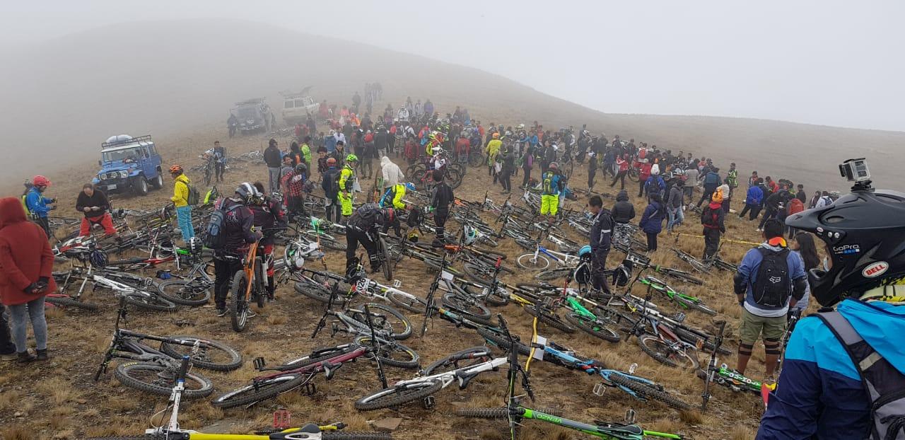 A large group of people is gathered on a foggy hillside, surrounded by numerous bicycles. Many individuals are wearing colorful biking gear and helmets, while some are engaged in conversations or tending to their bikes. A couple of off-road vehicles are parked nearby. The atmosphere appears lively, with a sense of community among the cyclists in a remote outdoor setting. Loma Loma mountain bike trail.