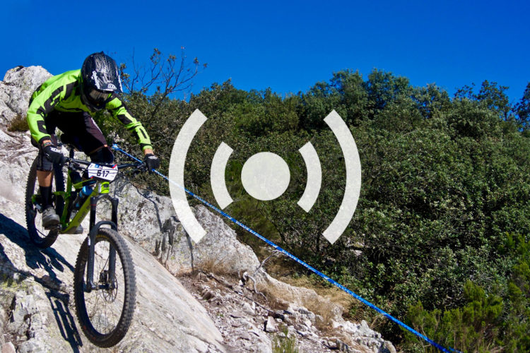 A mountain biker wearing a bright green jersey and protective gear navigates a rocky downhill trail. The sky is clear blue, and lush greenery is visible in the background. A blue rope is attached to the bike, suggesting an element of sport or competition.