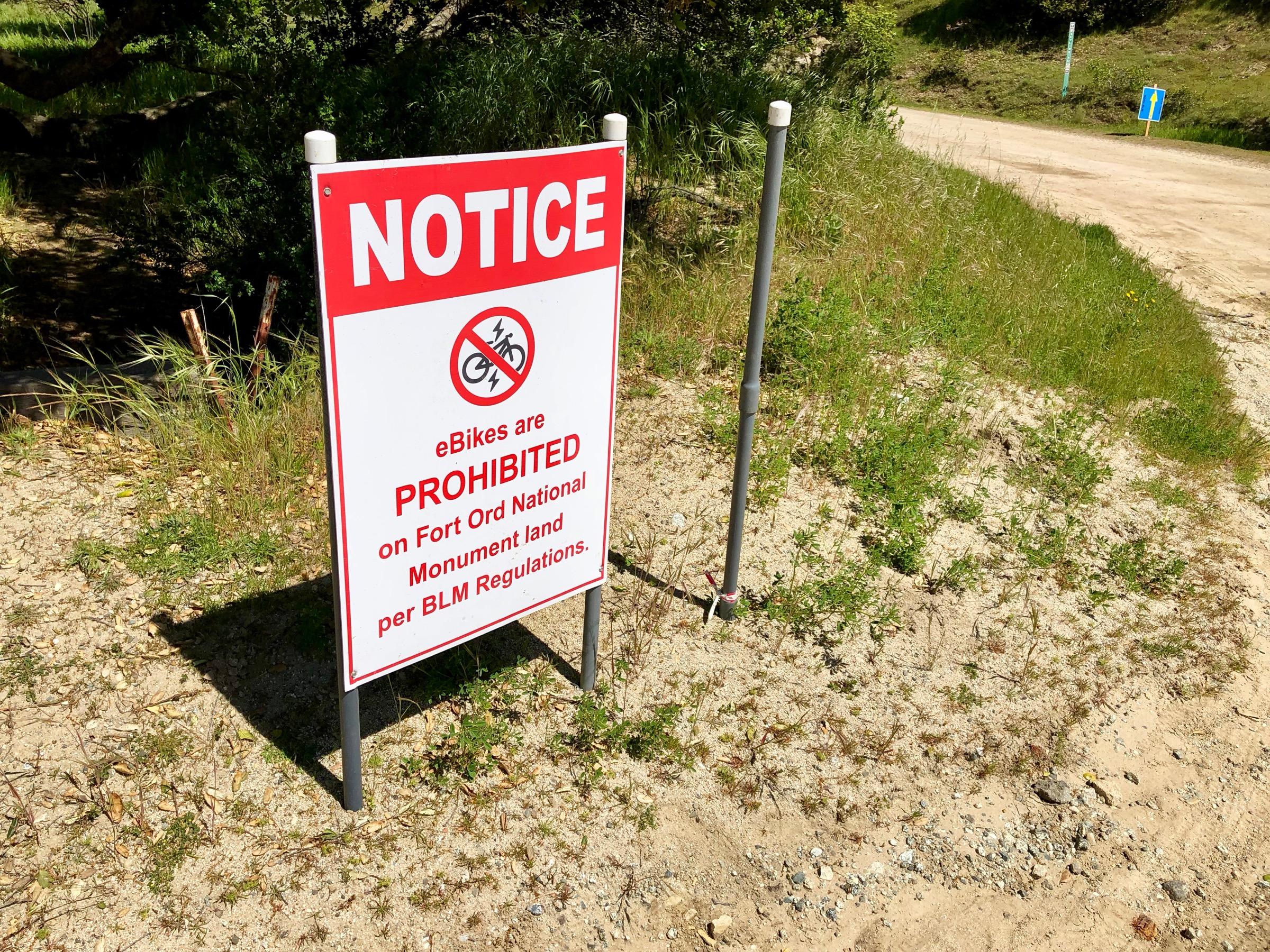 Sign indicating that eBikes are prohibited on Fort Ord National Monument land, with text reading "NOTICE" and "eBikes are PROHIBITED per BLM Regulations." The sign is situated in a grassy area near a dirt road. Fort Ord Public Lands mountain bike trail.