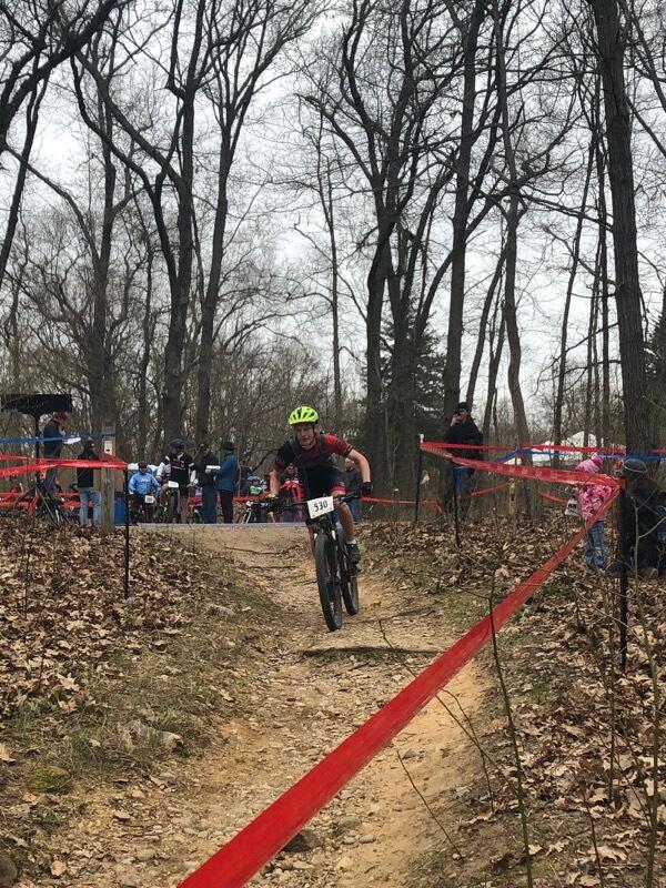 A young cyclist in a green helmet races down a dirt path marked with red tape, surrounded by trees. Spectators watch from the sidelines, while fallen leaves cover the ground. The scene captures the excitement of a mountain biking event on an overcast day. Yankee Springs mountain bike trail.