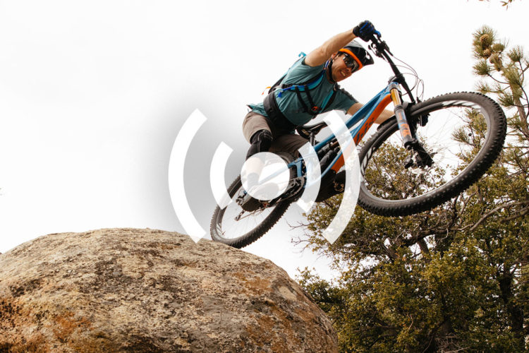 A mountain biker captured mid-air while jumping off a large rock, with trees in the background and a cloudy sky above. The rider is wearing protective gear and a helmet.