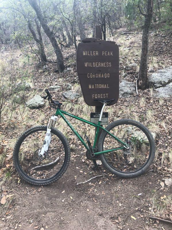 A green mountain bike is parked beside a rustic wooden sign that reads "Miller Peak Wilderness, Coronado National Forest." The sign is surrounded by a natural landscape of trees and rocky terrain, with scattered leaves on the ground. Brown Canyon mountain bike trail.