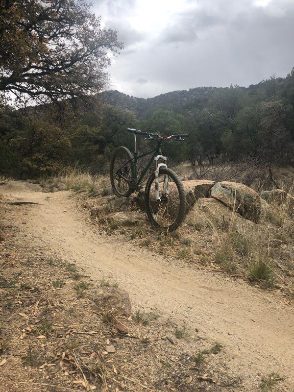 A mountain bike leaning against a rock on a dirt trail surrounded by sparse vegetation and hills under a cloudy sky. Brown Canyon mountain bike trail.