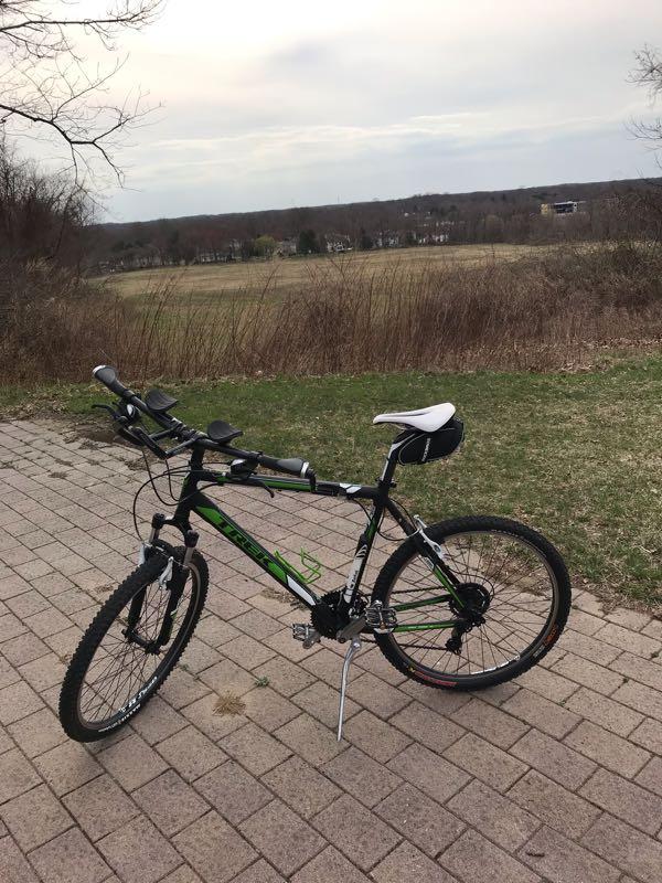 A green and black mountain bike is parked on a paved surface, with a scenic view of an open field and trees in the background. The sky is overcast, suggesting a cool day. Big Brook Park mountain bike trail.