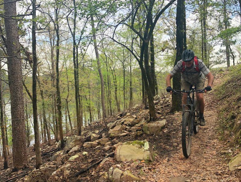 A mountain biker navigating a rocky trail surrounded by trees in a lush green forest, with a river visible in the background. Iron Mountain mountain bike trail.