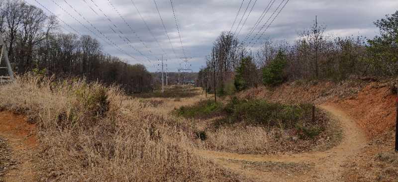A winding dirt path leads through a dry, grassy landscape under a cloudy sky, with power lines running overhead. Trees line the edges of the path, and a distant view of hills or buildings can be seen in the background. Wakefield mountain bike trail.