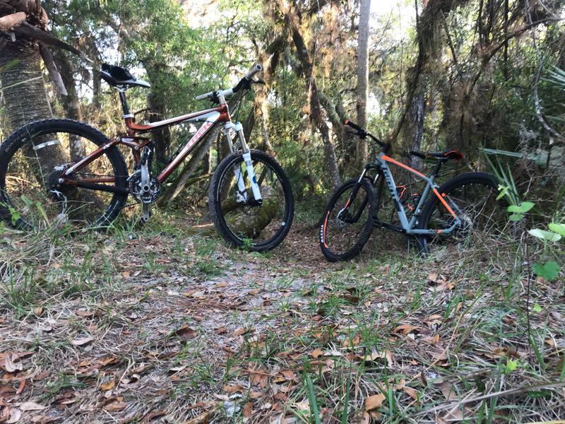 Two mountain bikes are parked on a narrow dirt path surrounded by lush green vegetation and trees. The bike on the left features a brown and white frame, while the one on the right has a gray and orange design. The scene conveys a sense of adventure and exploration in a natural setting. North Port Mountain Bike Trails mountain bike trail.