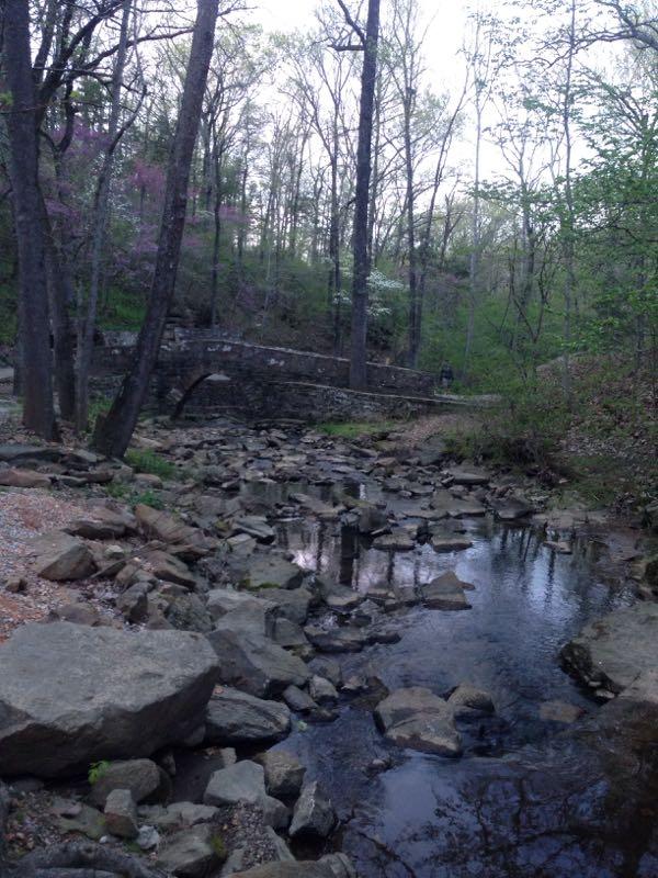 A serene view of a rocky stream flowing through a wooded area, surrounded by trees and scattered boulders. In the background, a stone arch bridge is visible, partially obscured by the trees. The scene is tranquil with hints of spring foliage and soft natural lighting. Mountain View City Park Trail mountain bike trail.