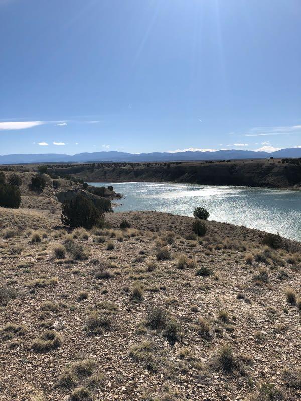 A scenic view of a river winding through a dry landscape, surrounded by gentle hills and sparse vegetation under a clear blue sky. The sunlight reflects off the water, creating glimmers. Low shrubs are visible on the riverbank, with distant mountains in the background. Voodoo mountain bike trail.