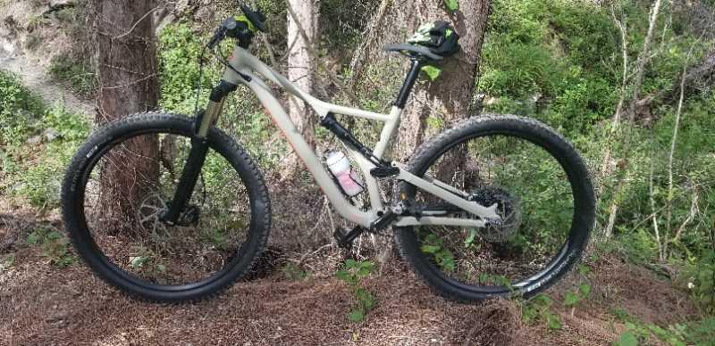 A mountain bike parked on a forest path, surrounded by trees and green foliage. The bike features a lightweight frame, thick tires, and a water bottle attached to the frame. Sunlight filters through the trees, highlighting the natural setting. Amelia Earhart Park mountain bike trail.