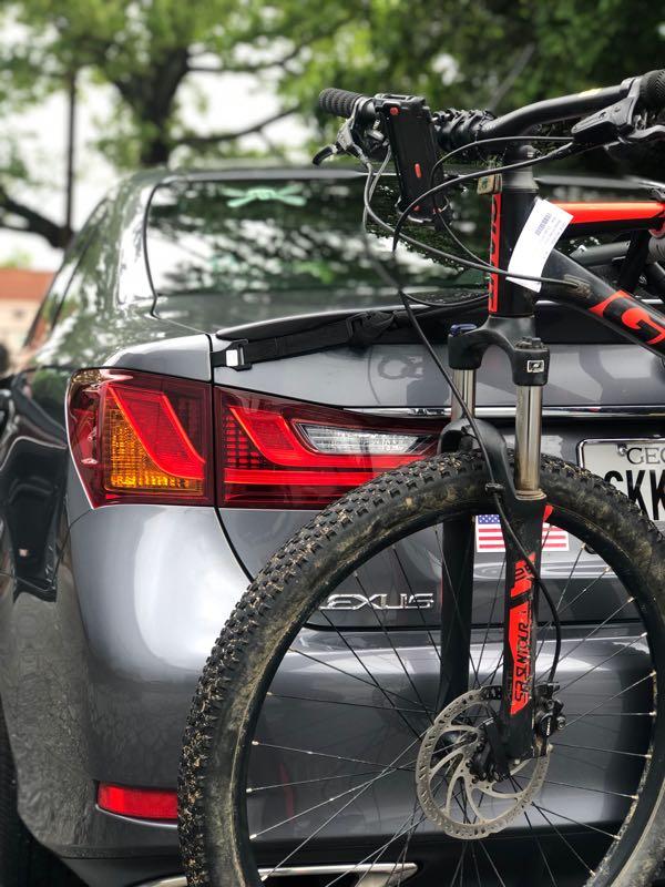 A gray Lexus vehicle is parked, displaying its rear and taillights. A mountain bike is securely mounted on a bike rack attached to the back, with a visible tire and brake disc. In the background, there are leafy trees, creating a natural setting.