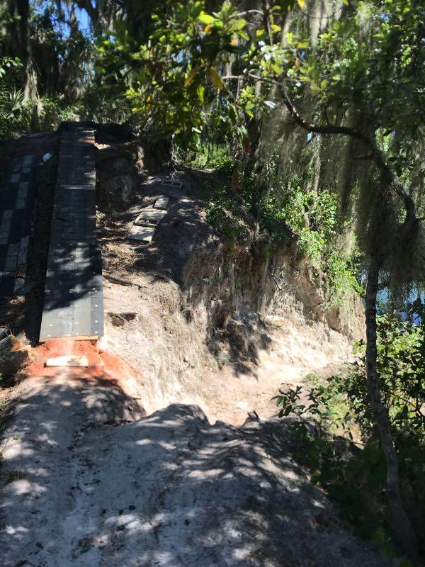 A wooden ramp leading down a sandy path surrounded by lush greenery, with a steep drop-off on one side and hints of water visible in the background, indicating a natural setting. The sunlight filters through the trees, creating dappled shadows on the ground. Loyce E. Harpe Park mountain bike trail.