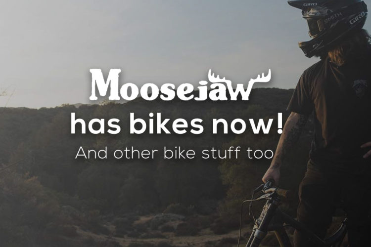 A mountain biker stands next to a bike in a natural outdoor setting, with trees and hills in the background. The image features overlay text that promotes Moosejaw's new bike offerings, stating "Moosejaw has bikes now! And other bike stuff too!"