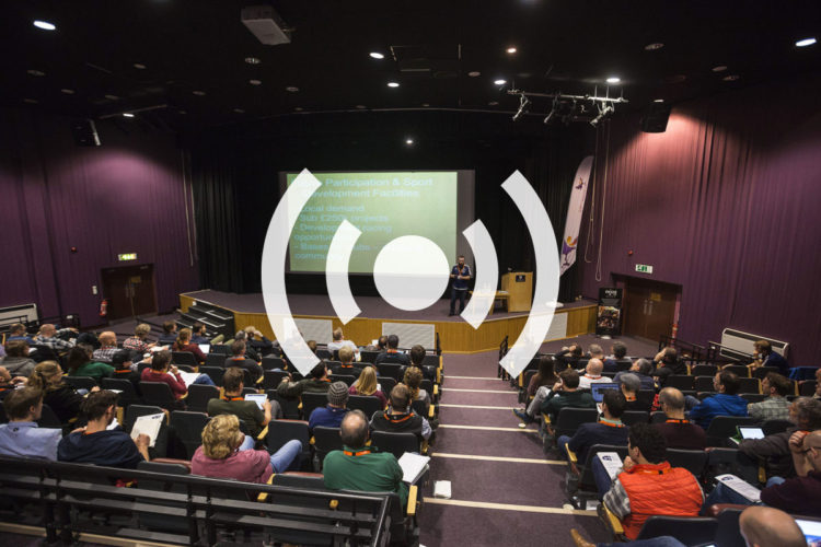 A speaker presents to an audience in a lecture hall. The audience, made up of various individuals seated in rows, appears to be engaged and taking notes. A presentation slide on the screen features text related to participation and sport development facilities. The setting includes purple curtains and a stage, creating a formal atmosphere for the event.
