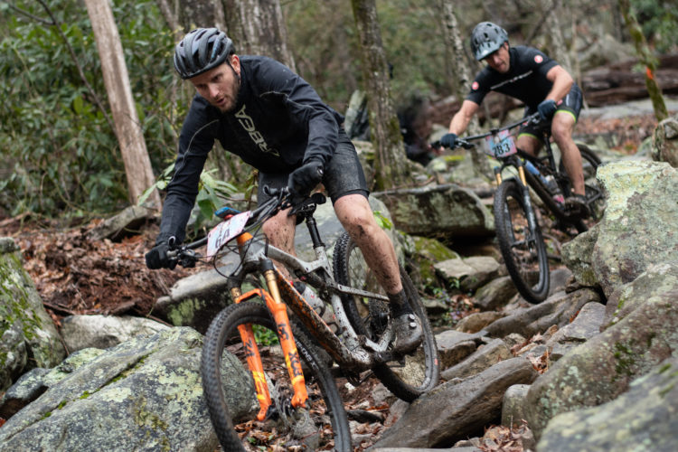 Two mountain bikers navigate a rocky, muddy terrain in a forested area. One rider leans forward, focused on maneuvering over the challenging rocks, while the other follows closely behind. Both riders are dressed in athletic gear and helmets, showcasing intense determination amidst the natural surroundings.