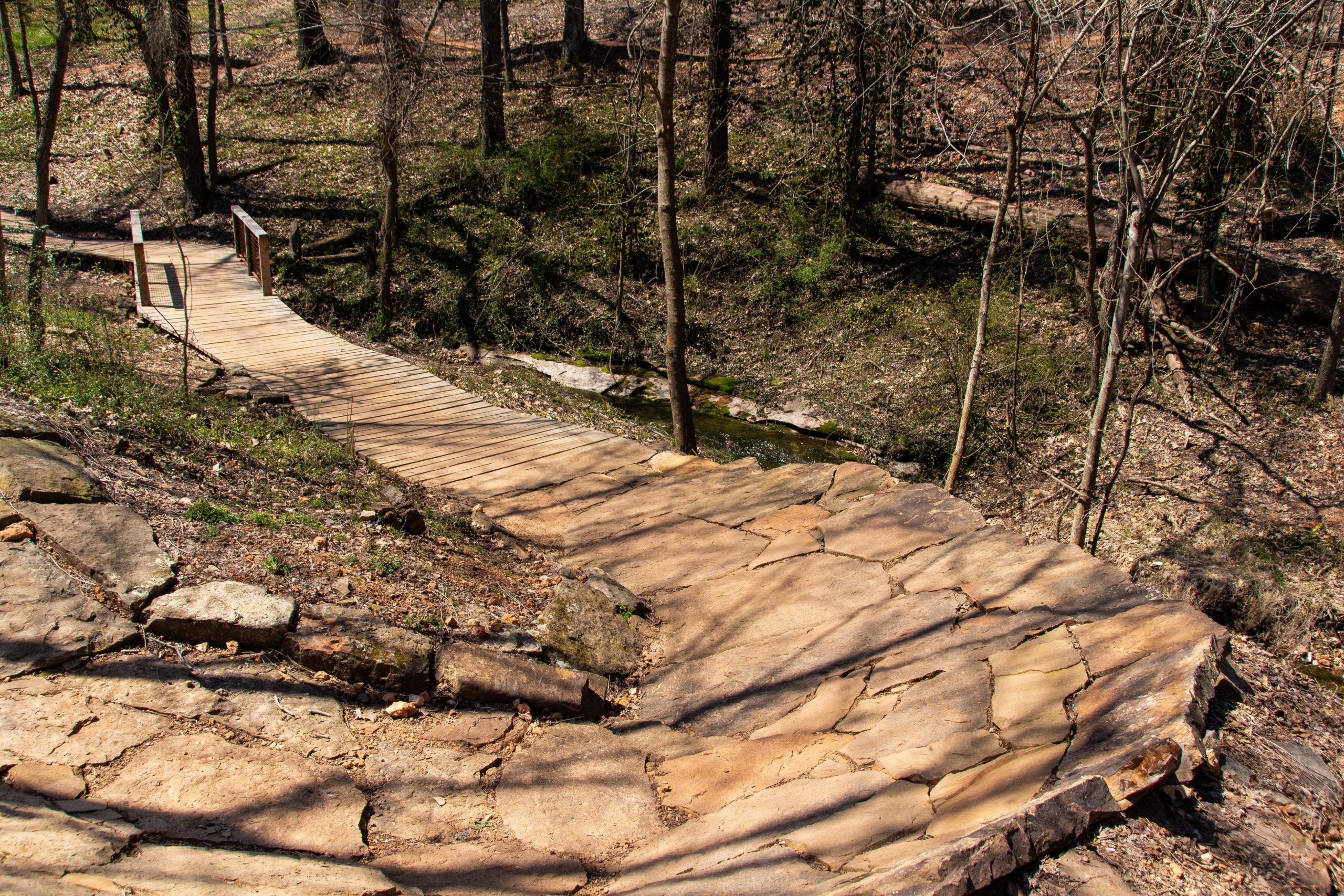 A winding wooden bridge crosses a small stream, surrounded by wooded terrain. The path leading to the bridge is made of large stone slabs, and the area is littered with fallen leaves, indicating early spring or late winter. Tall trees provide shade, creating a tranquil natural setting. Slaughter Pen Trail mountain bike trail.