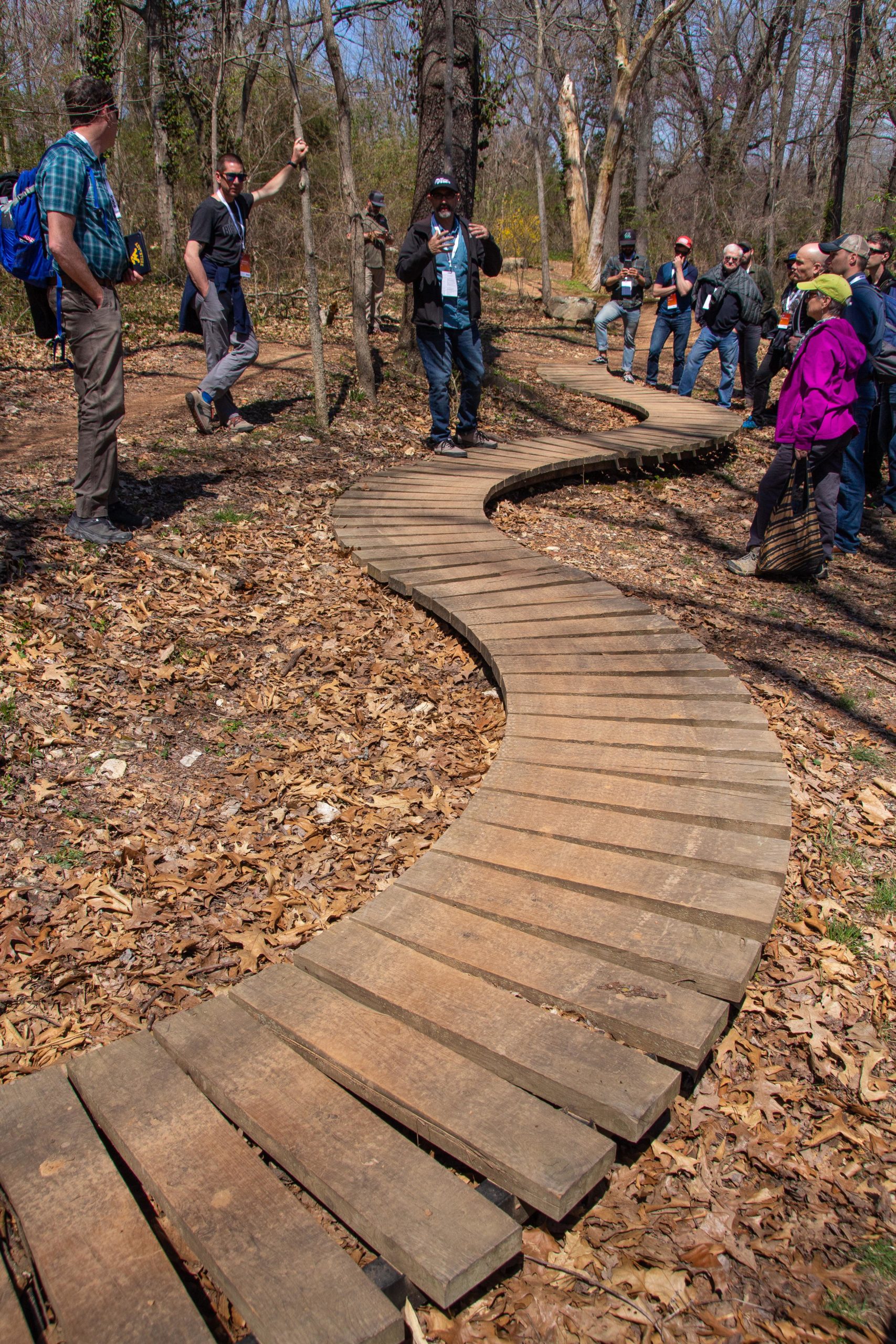 A group of people gathered in a wooded area, discussing on a curved wooden pathway surrounded by fallen leaves. The scene captures individuals in various outdoor clothing, with some leaning against trees and others listening attentively to a speaker. Sunlight filters through the trees, creating a serene natural setting. Slaughter Pen Trail mountain bike trail.