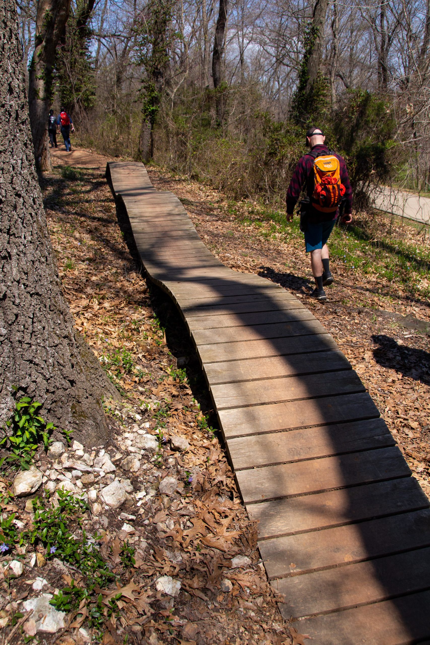 A winding wooden boardwalk pathway surrounded by trees and natural foliage, with two hikers in the distance. The ground is covered with fallen leaves, and the sunlight casts shadows on the boardwalk. Slaughter Pen Trail mountain bike trail.