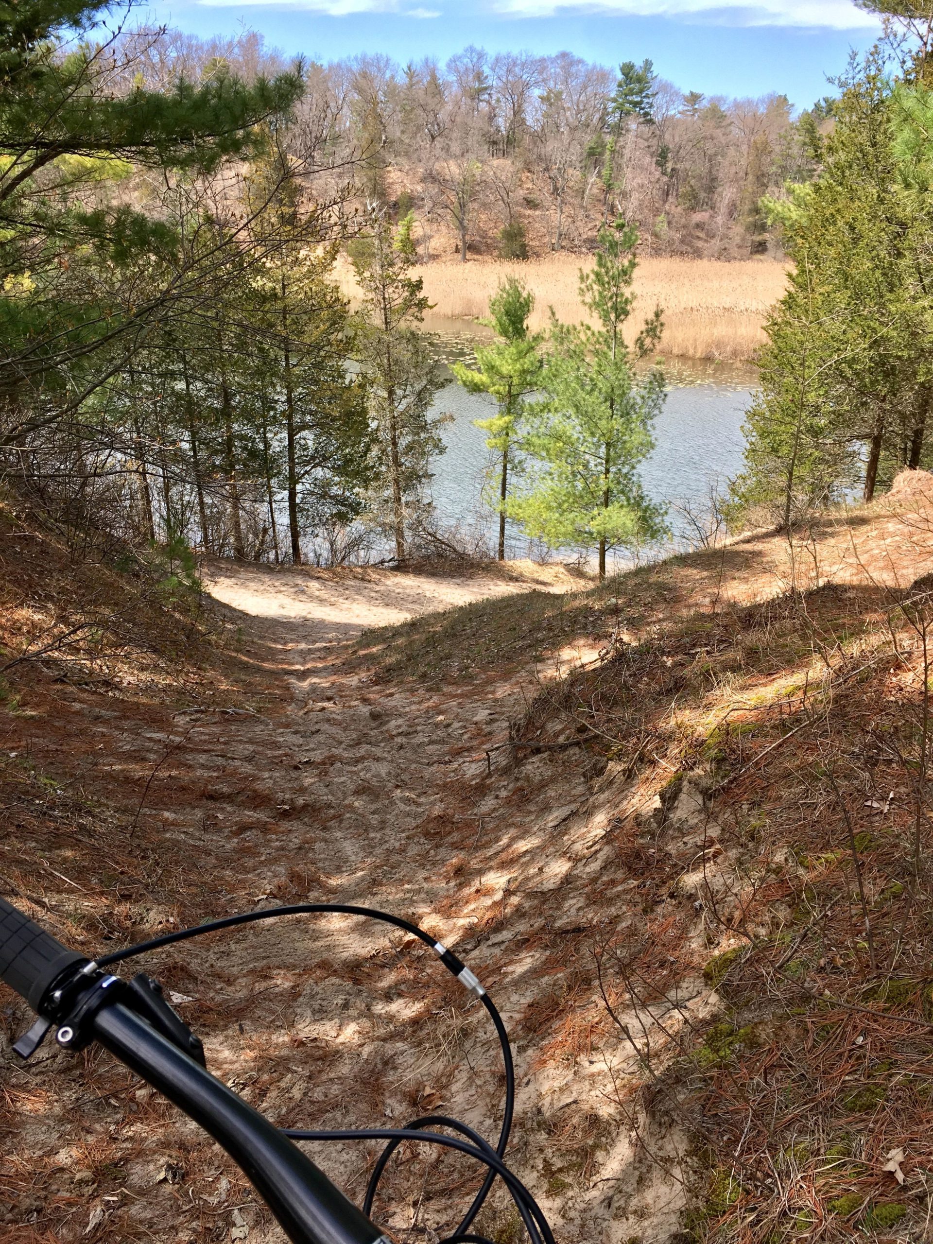 A view of a sandy pathway leading down toward a serene lake, framed by green pine trees and sparse foliage. The image captures the handlebars of a bicycle in the foreground, suggesting a biking trail. In the background, the lake reflects the blue sky and is bordered by tall grasses and wooded areas. Pinery Provincial Park mountain bike trail.
