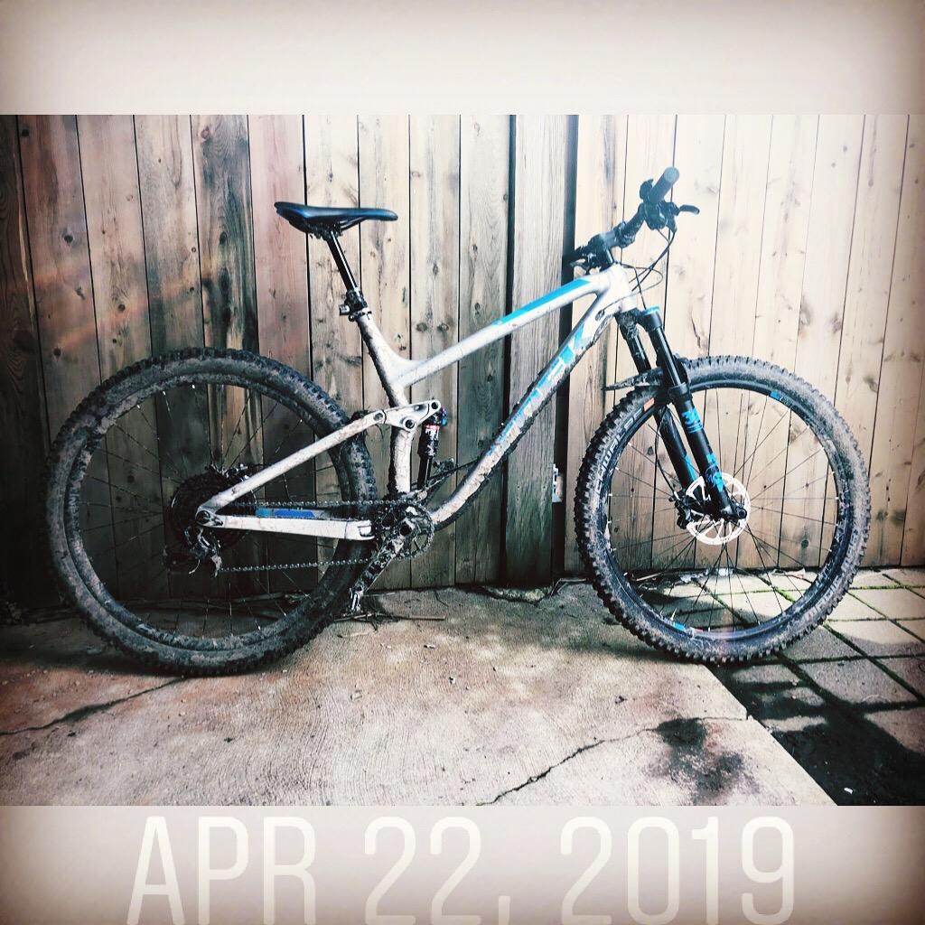Trek Fuel EX 9 29: A mountain bike with muddy tires rests against a wooden fence on a concrete surface, with the date "APR 22, 2019" displayed at the bottom of the image.