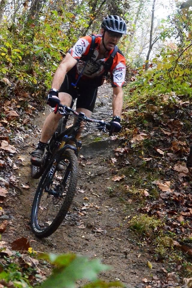 A mountain biker navigating a narrow, muddy trail surrounded by trees and fallen leaves. The cyclist is wearing a helmet and cycling gear, showcasing a concentrated expression as he rides over rough terrain. Angler's Ridge mountain bike trail.