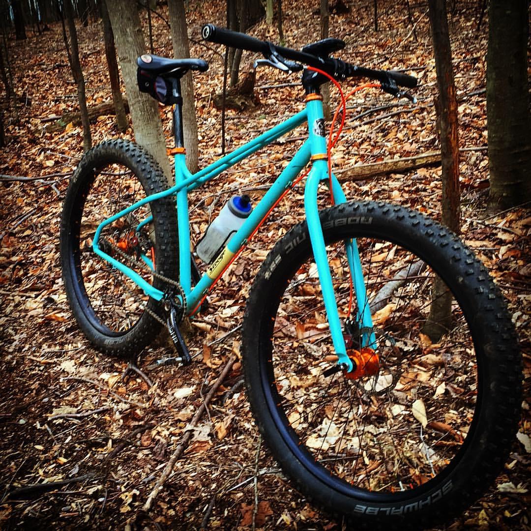 Stooge Stooge Mk2: A colorful mountain bike with a turquoise frame and orange accents, parked on a forest floor covered in brown leaves. A water bottle is attached to the bike's frame, and trees surround the scene.