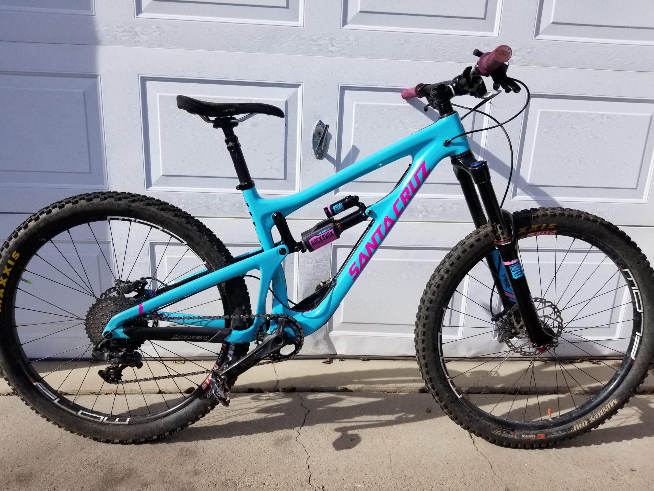 Santa Cruz Nomad: A bright blue mountain bike with pink accents, featuring a dual suspension, wide tires, and a dropper seat post. The bike is positioned against a white garage door, showcasing its detailed components including a rear derailleur and suspension forks.