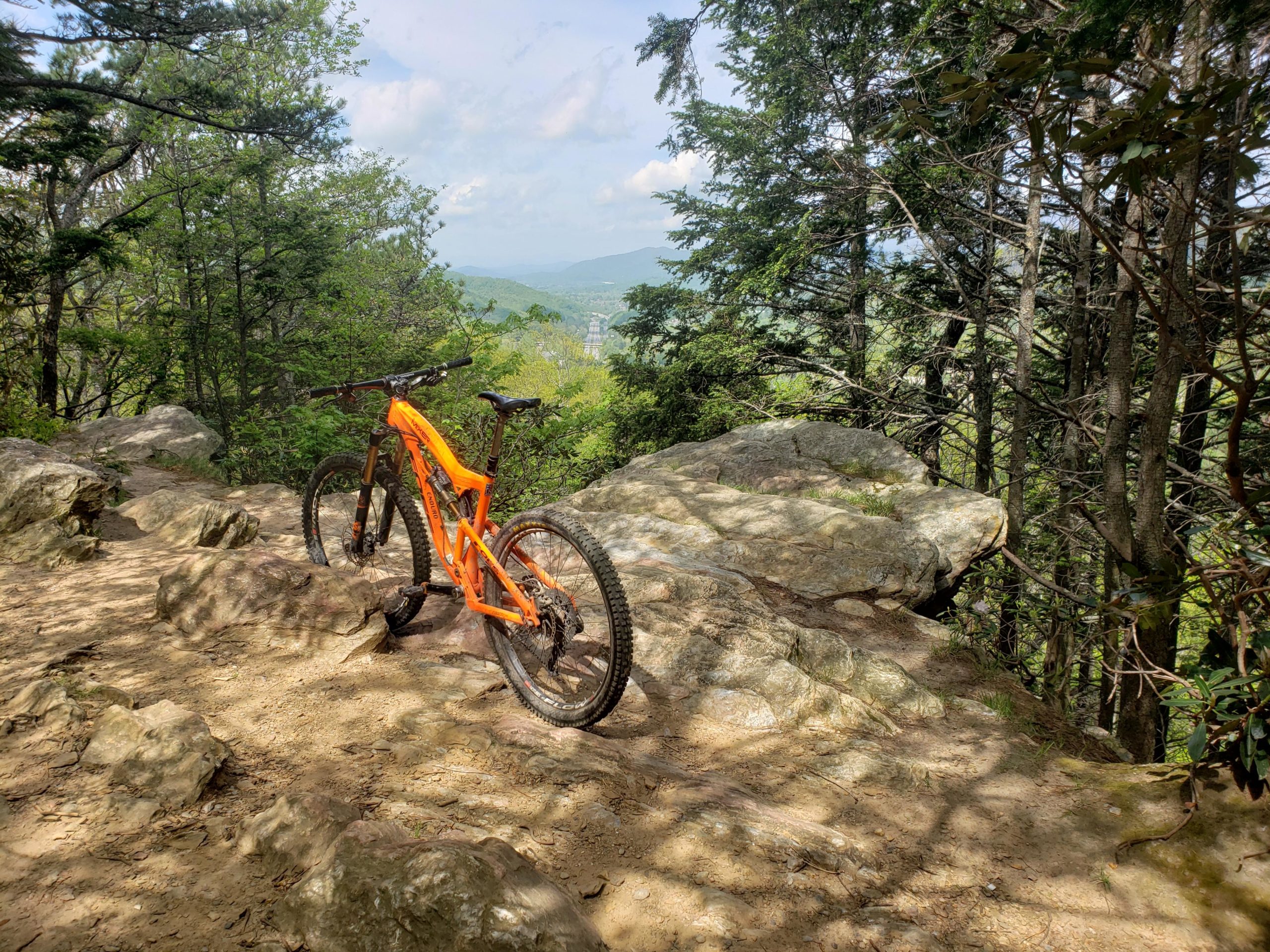 A bright orange mountain bike resting on a rocky trail surrounded by trees and greenery, with a scenic view of distant mountains in the background under a partly cloudy sky. Kitsuma mountain bike trail.