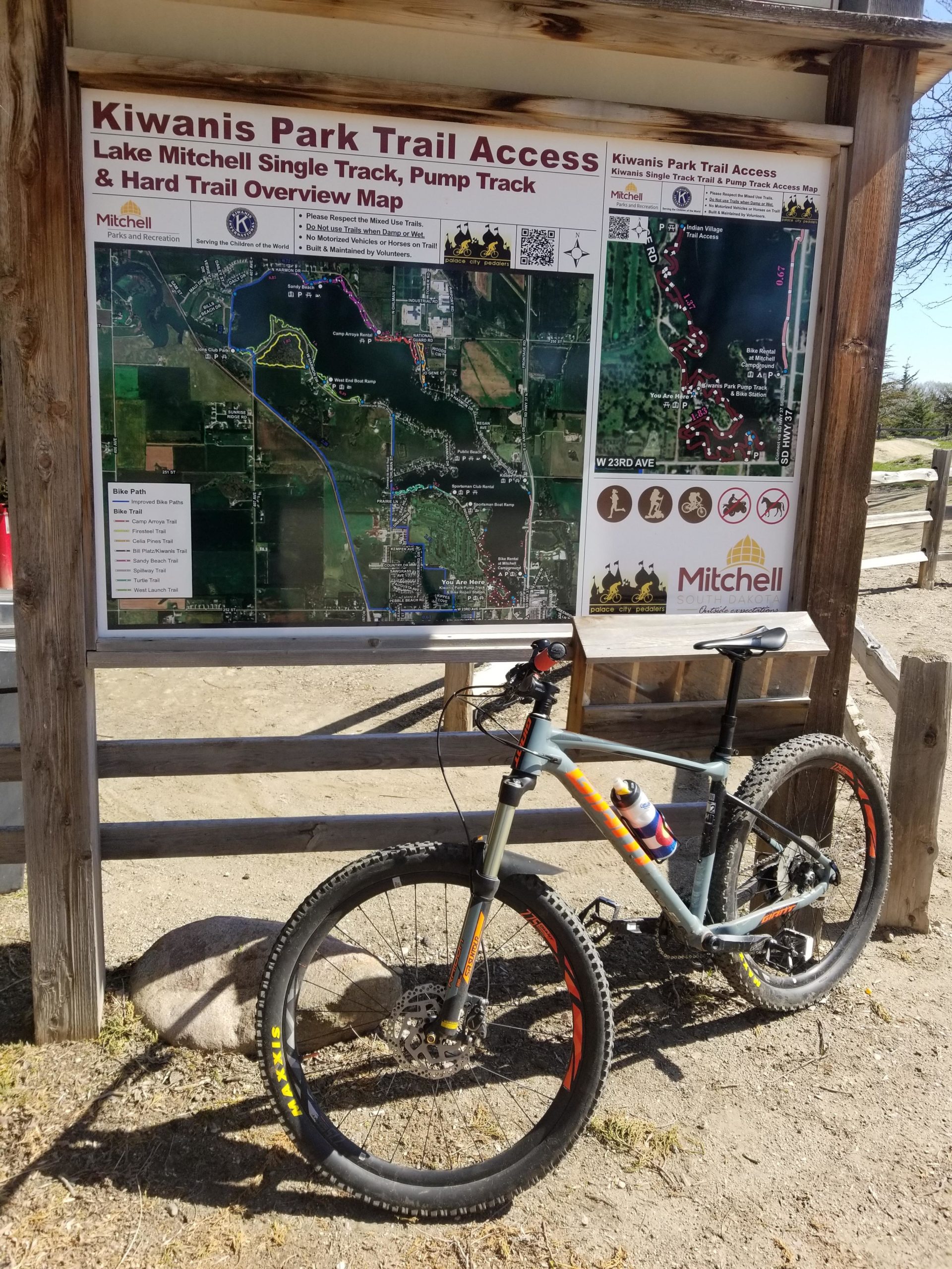 A mountain bike is parked next to a trail map displaying the Kiwanis Park Trail Access, which includes routes for the Lake Mitchell Single Track, Pump Track, and Hard Trail. The map shows various trails and features around Lake Mitchell, along with a clear layout of the surrounding area. Kiwanis mountain bike trail.