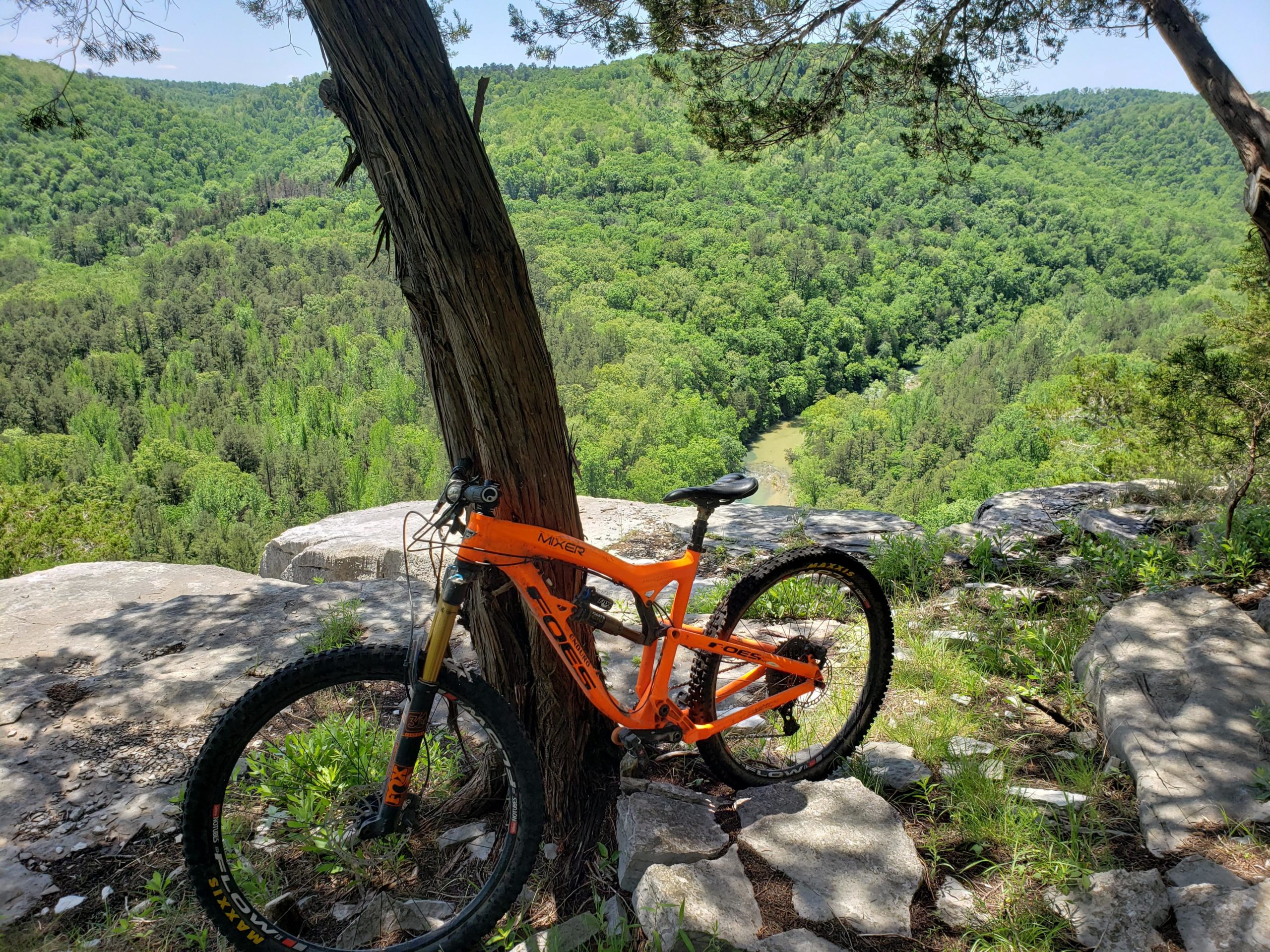 A bright orange mountain bike leaning against a tree on a rocky overlook, with a lush green valley and river visible in the background. The scene captures a sunny day with clear blue skies and vibrant foliage. Syllamo Trails mountain bike trail.