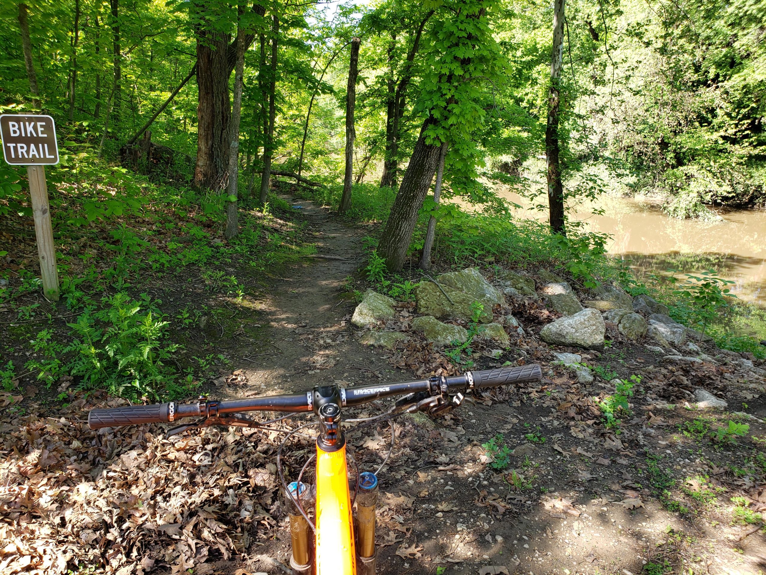 A view from the handlebars of a mountain bike on a dirt trail surrounded by green foliage, leading towards a calm river. A sign marking the area as a "Bike Trail" is visible on the left side of the image. Gunn Park Trails mountain bike trail.