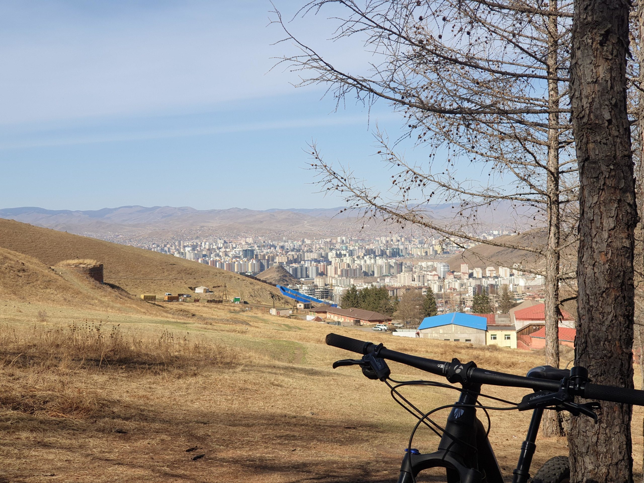 A mountain bike resting on a grassy hillside, framed by a tree, overlooking a sprawling cityscape with numerous buildings against a backdrop of distant mountains and a clear sky. Zaisan Chutes mountain bike trail.