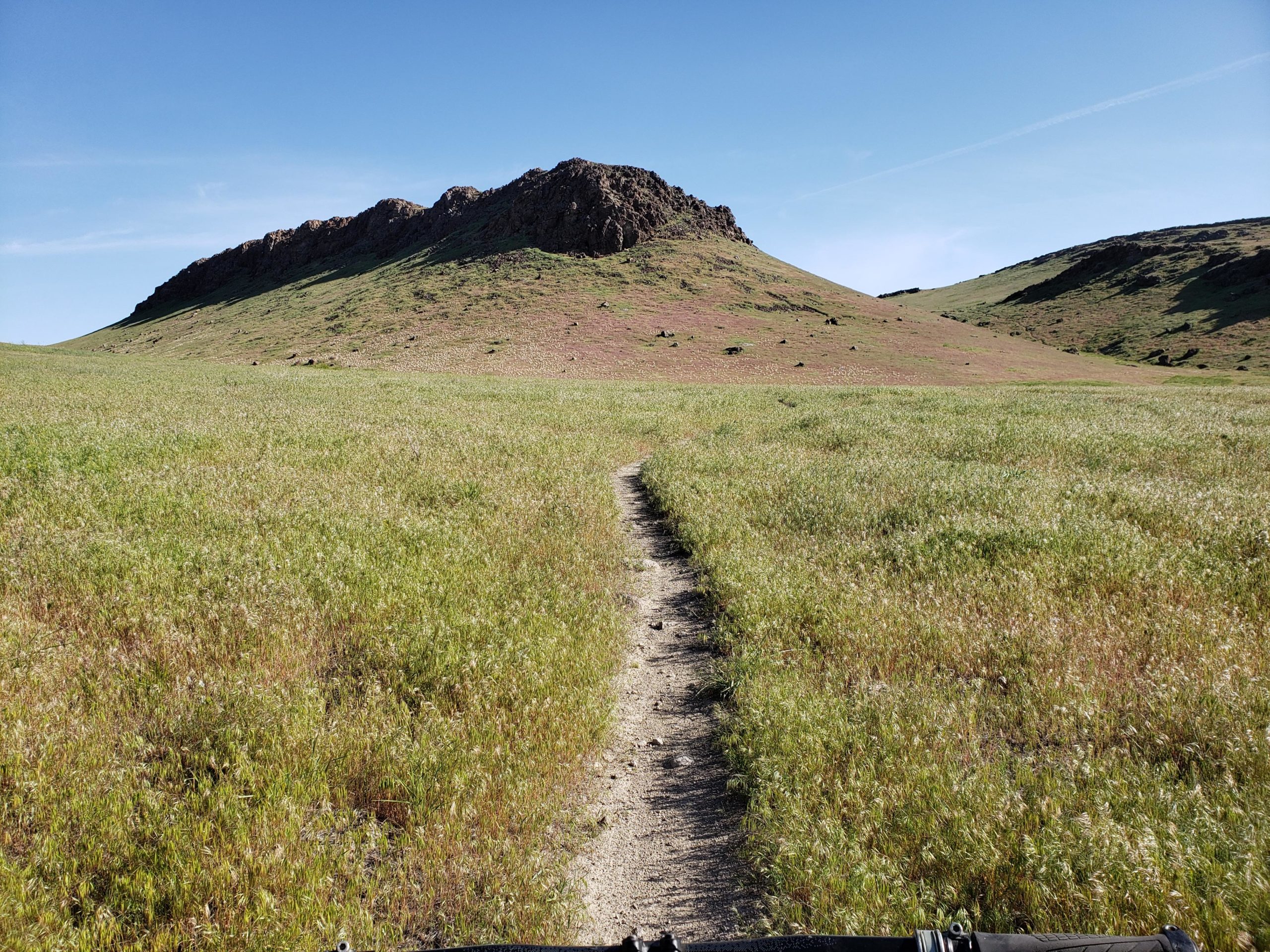 A dirt path leads through a grassy field towards a hill with rocky outcrops under a clear blue sky. The landscape is lush and green, with the hill rising gently in the background. Wilson Creek Area mountain bike trail.