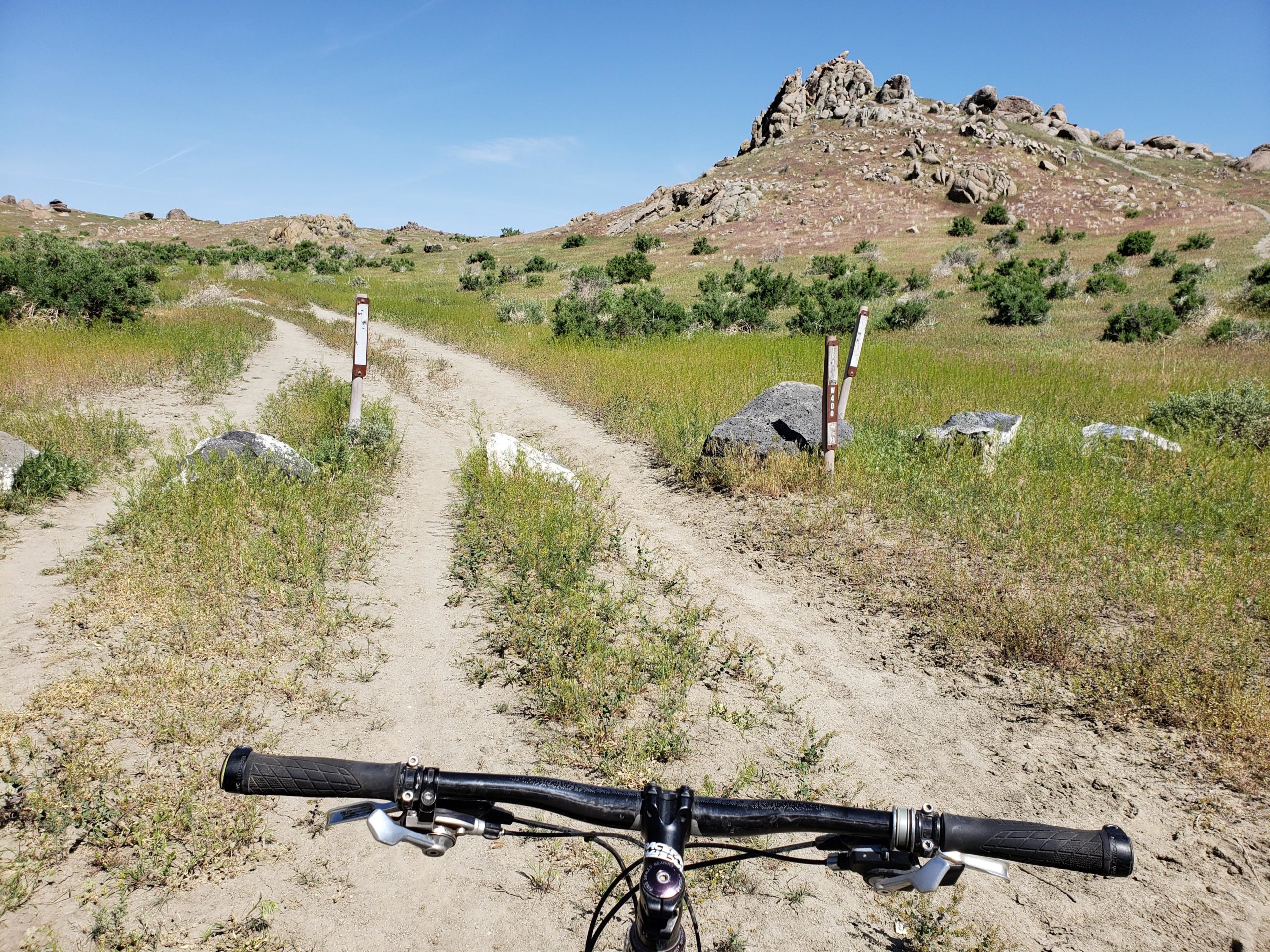 A mountain biker's perspective on a dirt trail winding through grassy terrain, with rocky hills in the background. Two trail markers are visible on either side of the path. The scene is set under a clear blue sky. Wilson Creek Area mountain bike trail.