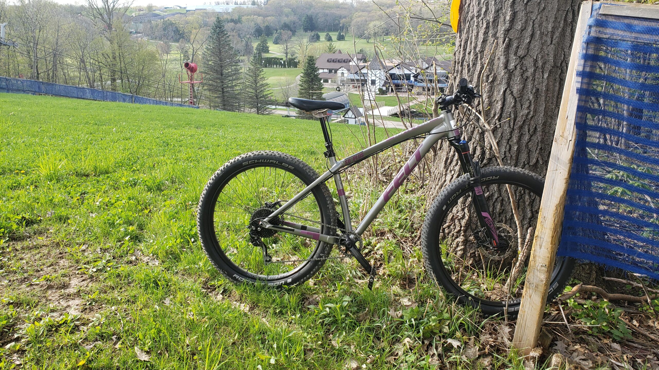 Felt Surplus: A mountain bike resting against a tree in a grassy area, with scenic hills and structures in the background. The bike features thick tires and a lightweight frame, surrounded by green grass and sparse trees.