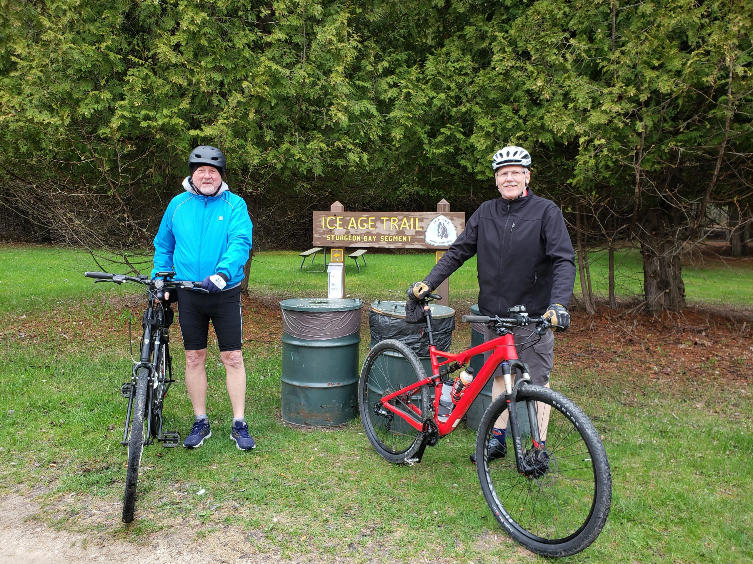 Two cyclists pose with their bikes near a sign for the Ice Age Trail, surrounded by greenery. One cyclist wears a blue jacket and black shorts, while the other is dressed in a black jacket and gray pants. A trash can is nearby, and picnic tables can be seen in the background. Ahnapee State Trail mountain bike trail.