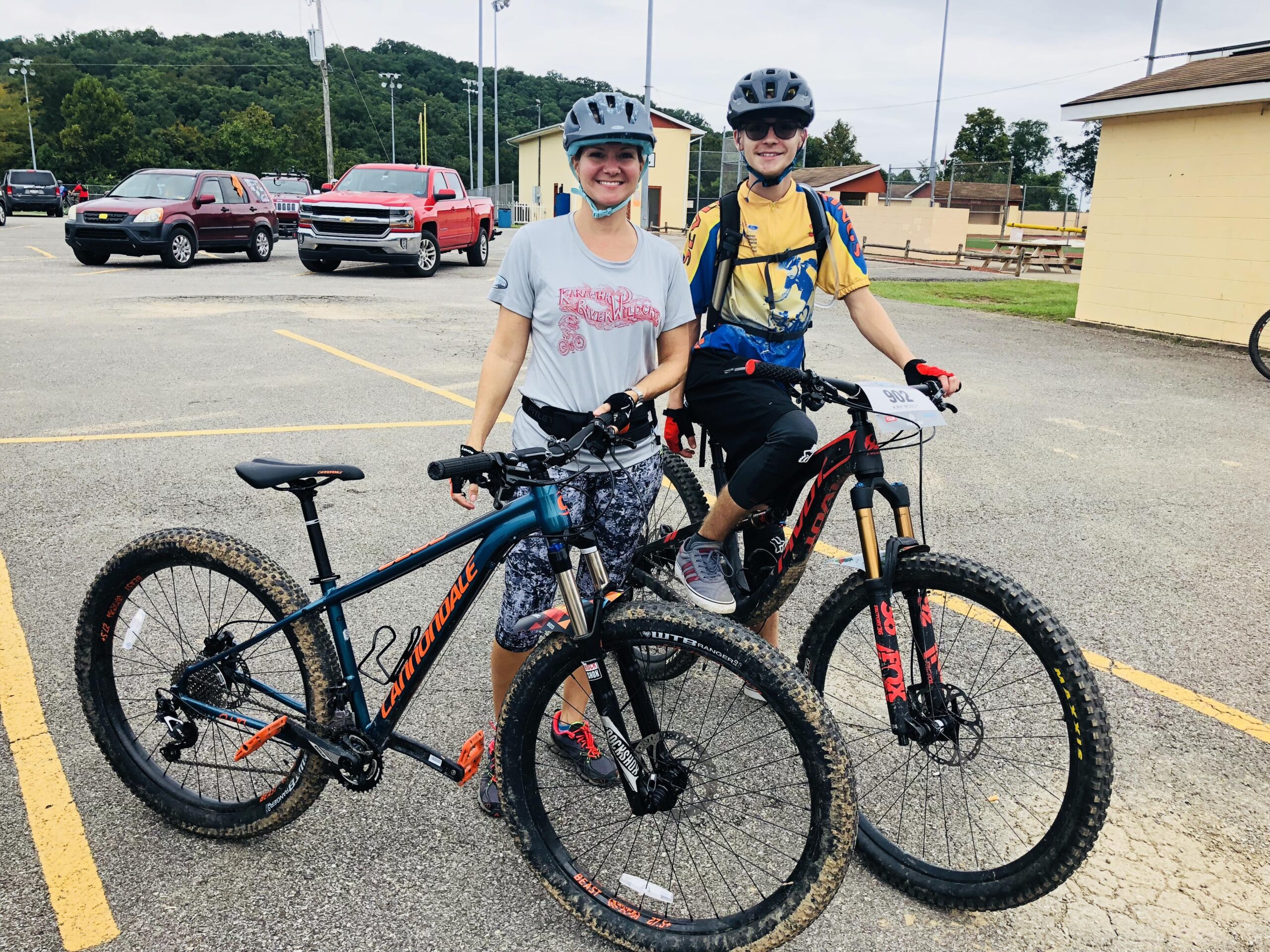 Cannondale Cujo 2: A woman and a young man stand next to their mountain bikes in a parking lot. The woman is wearing a gray t-shirt and patterned shorts, while the young man is dressed in a yellow and blue shirt with black shorts. Both are wearing helmets and are smiling at the camera. In the background, there are a few vehicles parked and a grassy area with buildings. The sky is overcast.