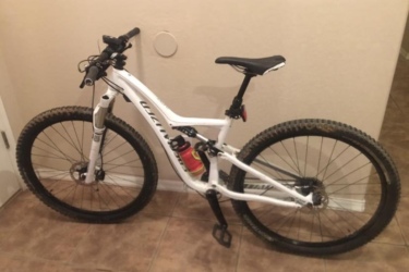 Specialized Rumor: A white mountain bike leaning against a wall in a tiled hallway. The bike features thick, knobby tires, a suspension fork, and a water bottle mounted in a cage on the frame.