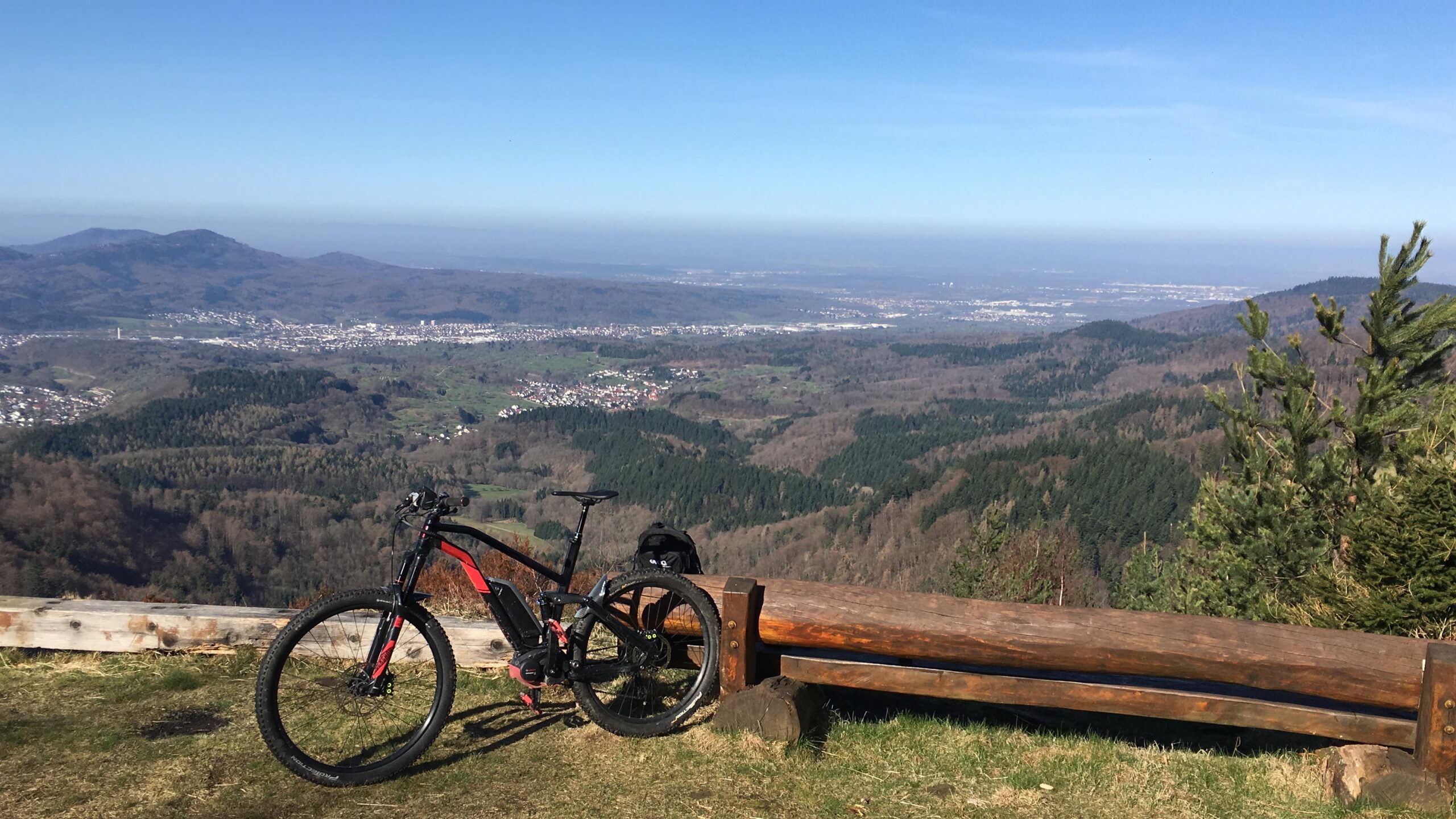 Moustache Samedi Race 5: A mountain bike rests against a wooden bench, overlooking a panoramic view of a valley filled with green hills and a small town in the distance. In the background, there are more mountains under a clear blue sky. The scene captures the beauty of nature and the thrill of outdoor cycling.