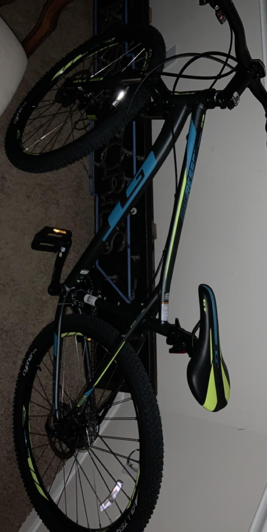 GT Aggressor Pro Mountain Bike: A black and blue mountain bike with green accents, laying on its side on a carpeted floor. The bike features thick tires suitable for off-road riding, a comfortable saddle, and a mounted front light. In the background, part of a chair and a wall can be seen, hinting at an indoor setting.