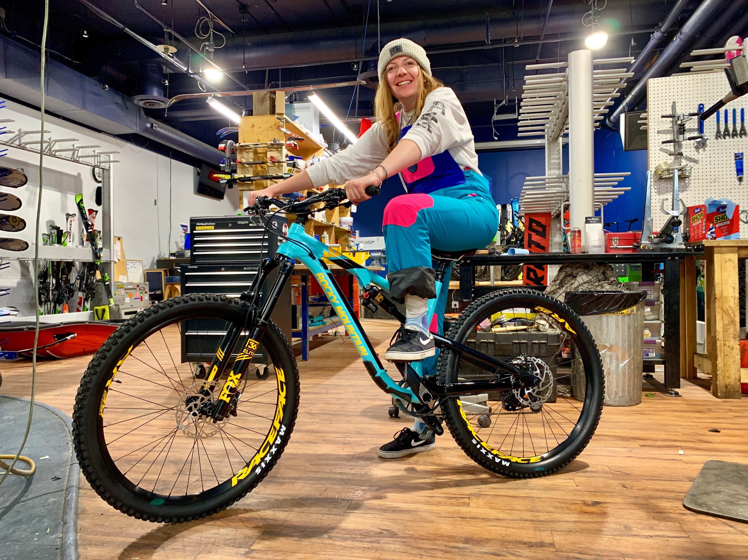 Rocky Mountain Altitude: A smiling person wearing a colorful outfit sits on a mountain bike inside a shop filled with various outdoor equipment and tools. The background includes a workbench with tools and a wall featuring sporting goods. The scene conveys an atmosphere of outdoor adventure and cycling enthusiasm.