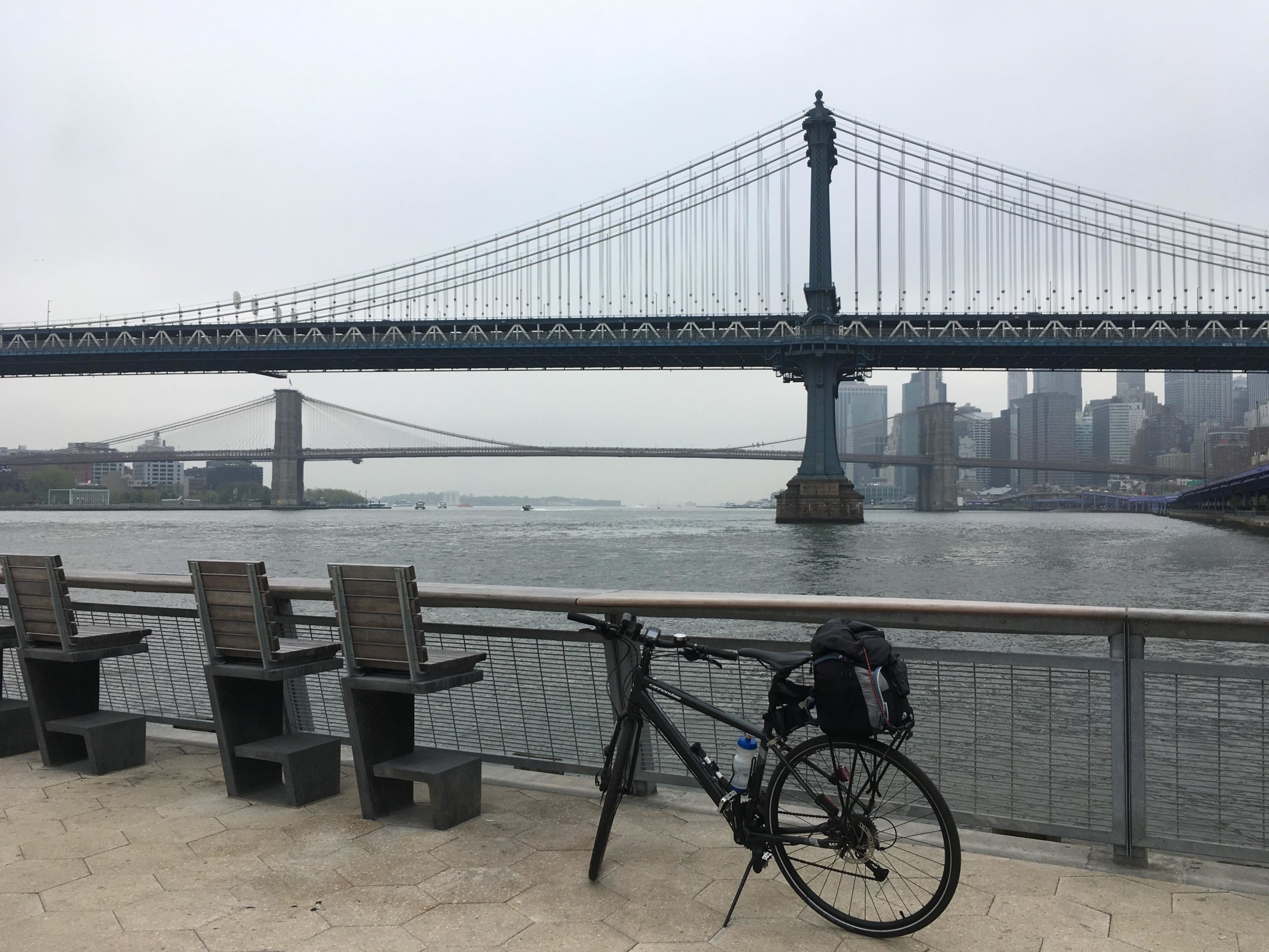 A bicycle rests beside a railing along a waterfront with a view of two bridges spanning the river. The scene is set on a cloudy day, with the city skyline visible in the background and empty wooden seats facing the water. East Side Green way 34th st to the Staten Island Ferry mountain bike trail.