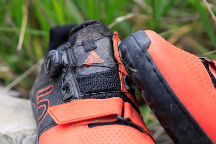 A close-up image of a pair of black and orange cycling shoes, featuring a lace system and Velcro strap, resting on a wooden surface with green grass in the background.