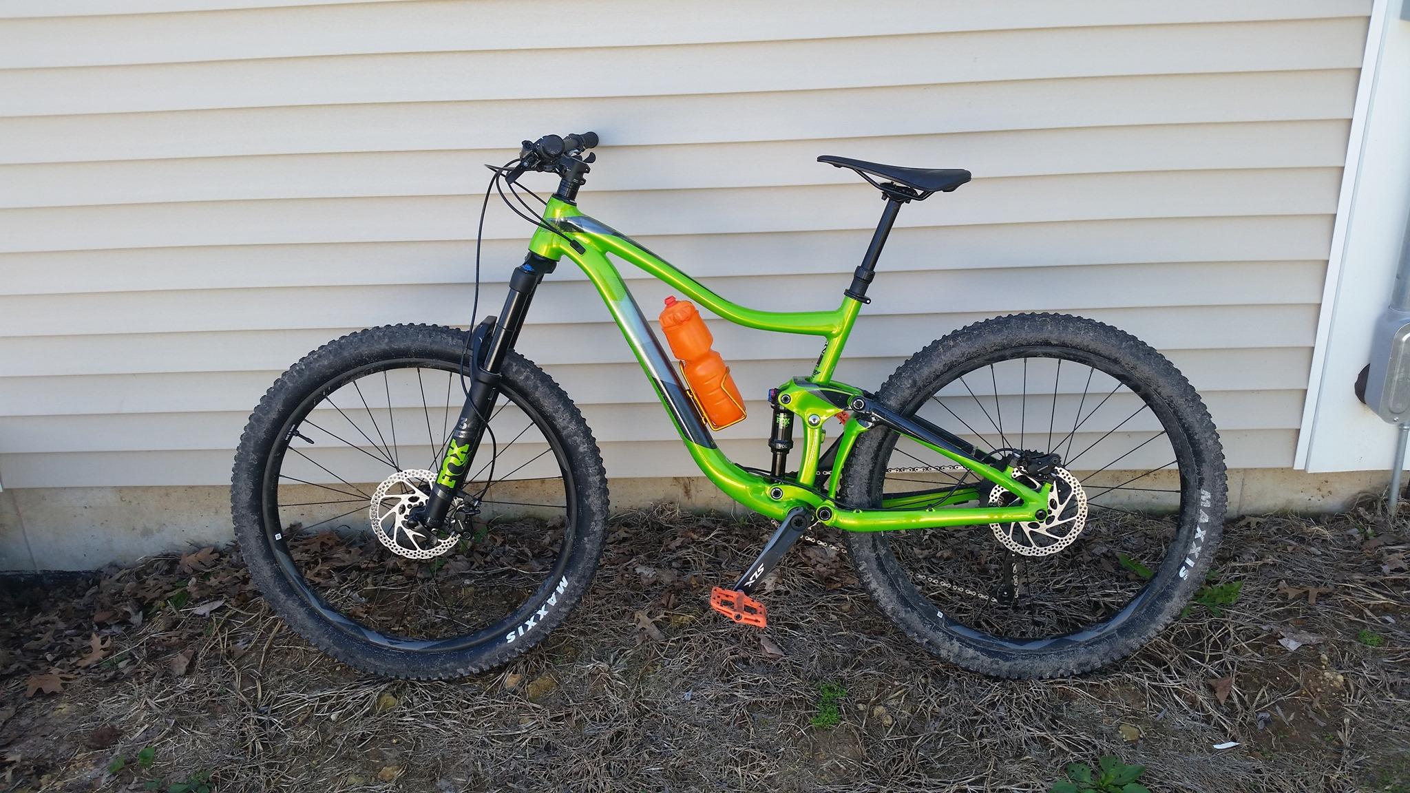 Giant Trance 27.5 2: A bright green mountain bike with black components, featuring wide tires, disc brakes, and an orange water bottle mounted in the frame. The bike is leaning against a beige vertical siding wall, with some dry leaves and grass visible on the ground.