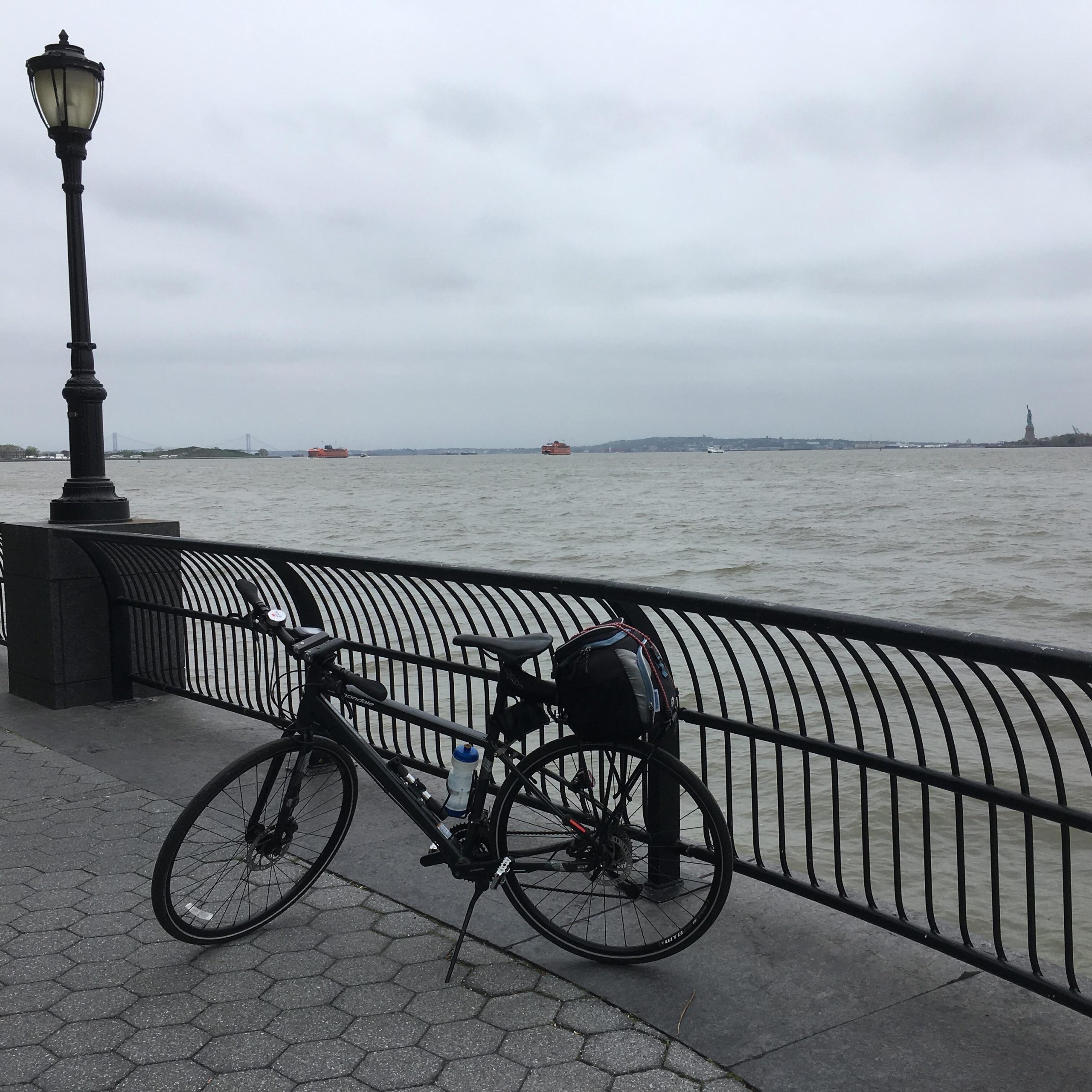 A black bicycle parked along a waterfront pathway, with a water view under a cloudy sky. Nearby, a lamppost is visible, and the Statue of Liberty can be seen in the distance, along with two orange boats on the water. West Street Greenway mountain bike trail.