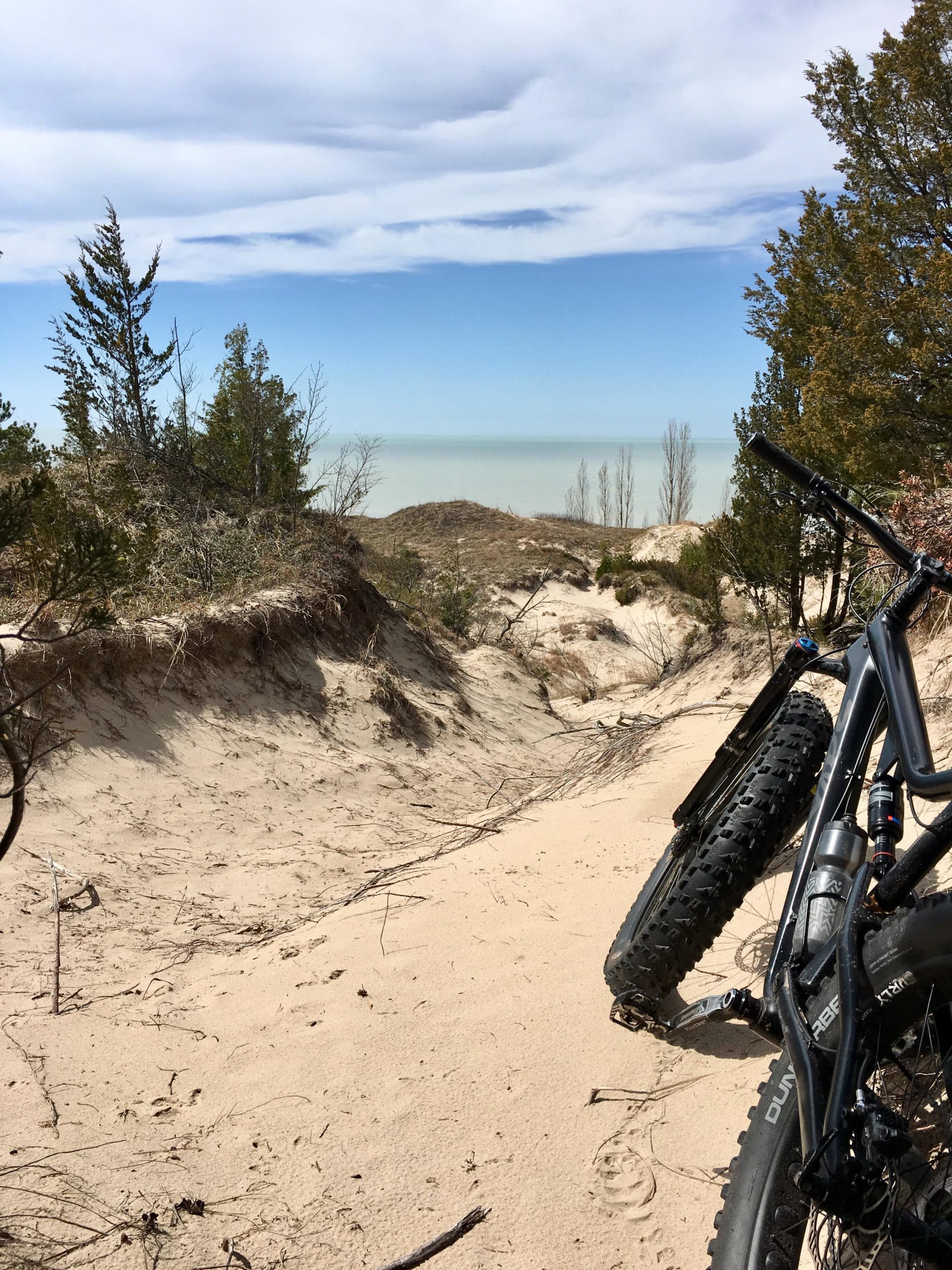 A sandy bike trail leading towards a view of the lake, with a bicycle leaning against a small dune. Sparse trees are visible on either side, and the sky is mostly clear with some clouds. Pinery Provincial Park mountain bike trail.