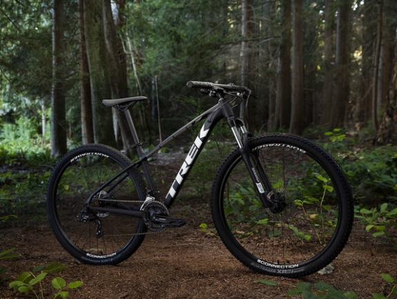Trek Marlin 5: A black mountain bike with the brand name "TREK" prominently displayed, positioned on a bed of soft earth surrounded by tall trees and greenery. The bike is angled to showcase its frame and tires, with a natural forest backdrop creating a rugged outdoor setting.